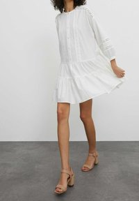 Robe blanche jusqu'aux genoux avec manches longues, boutonnée à l'avant, détails en dentelle et jupe à volants. Associée à des sandales beiges à talons blocs.