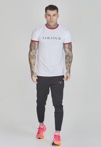 Witte t-shirt met rode rand en zwart "SIKSILK" logo, gecombineerd met zwarte joggers met een klein logo en helderroze sportschoenen.