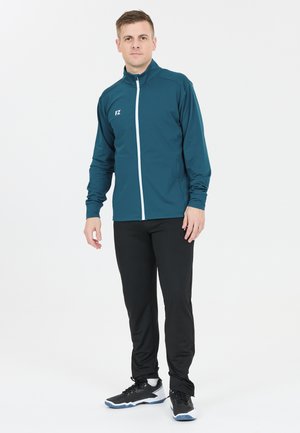 Mand står iført en teal zip-up jakke med "FZ" logo, sorte bukser og sorte og hvide sportssko på en hvid baggrund.