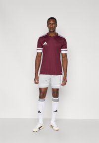Dunkelrote Sporttrikot mit weißen gestreiften Ärmeln, kombiniert mit hellgrauen Shorts. Weiße Socken mit schwarzen Streifen und Logo, goldene Fußballschuhe.