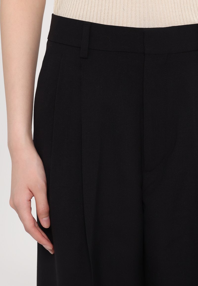 Black tailored trousers with a high waist, featuring pleats, side pockets, and a smooth fabric texture. Close-up on waist and hand.