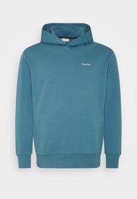 Sudadera de color azul verde de Calvin Klein con bolsillos frontales, puños y dobladillo acanalados, y pequeño logo blanco en el lado izquierdo del pecho.
