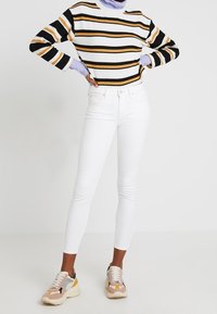 Suéter de rayas acanaladas en blanco, negro y amarillo; jeans ajustados blancos; zapatillas gruesas multicolores con una mezcla de tonos pastel.