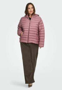 Kvinne iført en lilla, quiltet hettejakke med frontlommer, brune vide cordbukser og brune spisse sko, stående på hvit bakgrunn.