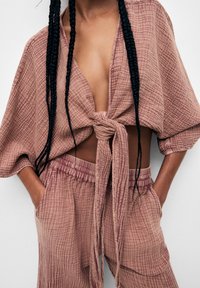 Femme portant un ensemble assorti texturé couleur vieux rose, avec un haut à manches larges noué devant et un pantalon ample, mains dans les poches, et cheveux longs tressés.