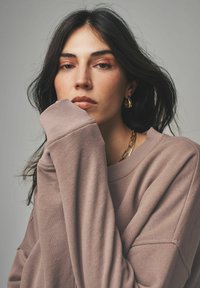 Sweatshirt i mjukt mauve-tyg, med ribbade ärmslut, vid båthalsringning, som bärs avslappnat med en hand lyft. Guldhalsband och hoop-örhängen.