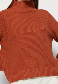 Sweter w kolorze rdzawego brązu, o fakturowanym splocie, z wysokim, żebrowanym kołnierzem i żebrowanym dołem. Posiada poziomy detal w postaci panelu na klatce piersiowej.
