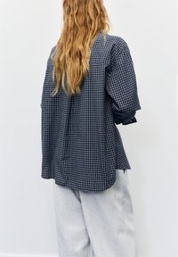 Navy checkered shirt with loose fit and dropped shoulders, featuring long sleeves and a curved hem, paired with light gray sweatpants.