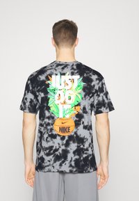 Hombre con camiseta Nike de tie-dye en negro y gris, con un diseño colorido de plantas y el texto "Just Do It" encima de un balón de baloncesto naranja en la parte posterior.