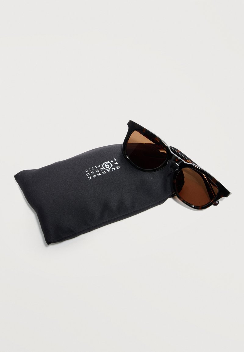 Occhiali da sole tartarugati con lenti scure poggiati su una custodia morbida nera con un logo numerico bianco su sfondo bianco.