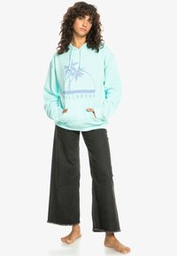 Licht turquoise hoodie met een grafische afbeelding van palmbomen en "BILLABONG" in het blauw. Gecombineerd met wijde zwarte denim broek en zonder schoenen gedragen.