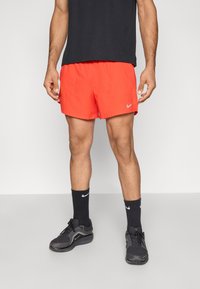 Κόκκινα αθλητικά σορτς με ελαστική μέση και πλαϊνές τσέπες, σε συνδυασμό με μαύρες κάλτσες και παπούτσια Nike, που στέκονται σε γκρι φόντο.