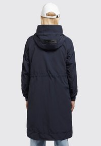 Marineblå lang pufferjakke med hætte, med indsnøret talje, ribbede manchetter og et sort logo-patch på ryggen. Glat, skinnende tekstur.