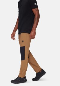 Tan outdoorbroek met zwarte kniestukken, voorzien van een logopatch en een relaxte pasvorm. Gepaarde met een zwart shirt met korte mouwen en zwarte schoenen.