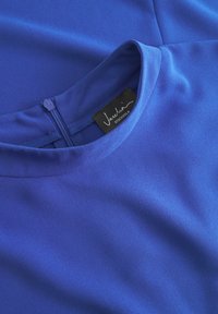 Modra majica iz gladkega materiala z okroglo ovratnikom, zadrgo na hrbtu in oznako z napisom "Jascha Stockholm." Prepoznaven videz in barva.