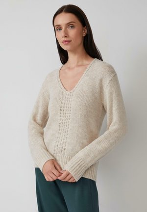 Frau mit glattem, dunklem Haar, die einen cremefarbenen V-Ausschnitt-Strickpullover und dunkelgrüne Hosen trägt und vor einem schlichten hellen Hintergrund steht.