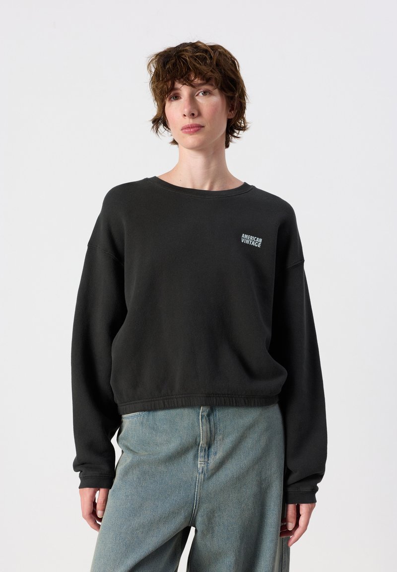 Persona joven con cabello corto y castaño que lleva una sudadera negra y vaqueros azul claro, de pie frente a un fondo blanco liso.