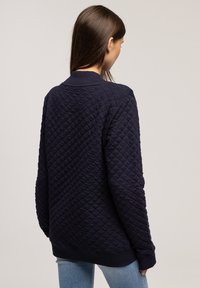 Granatowa pikowana bluza z teksturowanym wzorem, ściągaczem przy kołnierzu i mankietach. Tył ma luźny krój i prosty design.