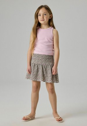 Mladé dievča stojí, oblečené v ružovom tielku bez rukávov, kvetinovej sukni a béžových žabkách na jednoduchom svetlosivom pozadí.