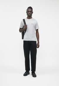 Szürke rövid ujjú póló piros szöveggel, fekete egyenes szárú nadrággal és fekete cipővel párosítva. A modell egy fekete táskát tart.