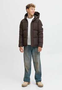 Giacca marrone imbottita con cappuccio, dotata di chiusura con zip e bottone. Indossata sopra un maglione bianco, abbinata a jeans in denim blu larghi e scarpe chiare.
