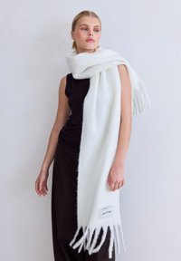 Witte wollen sjaal met franjes, met een zachte textuur en een label met de tekst "ARCTIC FOX", over een zwart bovenstuk gedrapeerd.