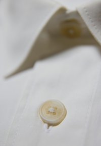 Close-up van een beige knop die is genaaid op de voorkant van de plooi van een witte overhemd dicht bij de kraag.