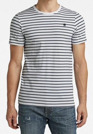 Mann trägt weißes Kurzarm-T-Shirt mit dünnen schwarzen horizontalen Streifen und kleinem Logo auf der Brust, kombiniert mit abgenutzten blauen Jeans.