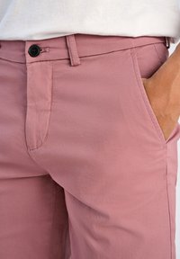 Pantaloni rosa chiaro con una texture di tessuto liscia, chiusura a bottone singolo e tasche laterali, con dettagli di cuciture curate.