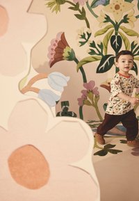 Barn iført en mønstret t-shirt med jordfarver og brune kordfløjlsbukser, gående på en lysfarvet gulv med blomsterdekorationer.