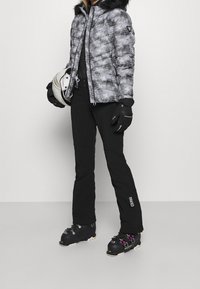 Padded czarna i biała kurtka w wzór z futrzanym kapturem, połączona z czarnymi spodniami narciarskimi i rękawicami. Wyposażona w buty narciarskie i kask.