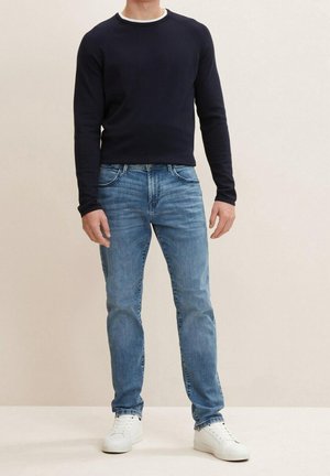 Mand iført sort langærmet skjorte, lyseblå jeans og hvide sneakers, stående mod en ensfarvet lys baggrund.