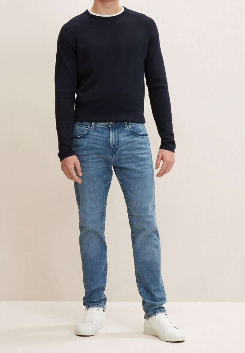 Homme portant un t-shirt noir à manches longues, un jean bleu clair et des baskets blanches, debout devant un fond clair uni.