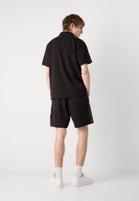 Čierne polo tričko a zodpovedajúce šortky vyrobené z priedušnej tkaniny, s jednoduchým dizajnom. Model má na sebe biele tenisky a krátke ponožky.