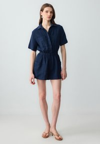 Combinaison courte en lin bleu marine avec manches courtes, fermeture à boutons, taille élastique et deux poches avant. Texture lisse et design décontracté.