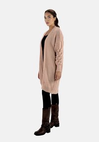 Ljusrosa stickad cardigan med texturerade ballongärmar, öppen front och ribbad nederkant, kombinerad med svarta leggings och bruna ankelstövlar.