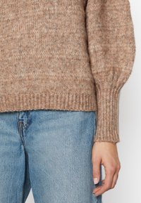 Pull en tricot beige doux avec un motif texturé, présentant des poignets et un ourlet côtelés, associé à un jean en denim bleu clair.