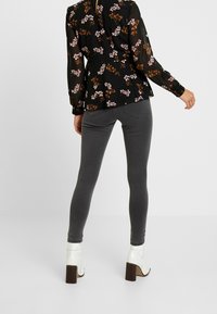 Schwarze Bluse mit floralem Muster und langen Ärmeln; kombiniert mit grauen Leggings und weißen Ankle-Boots mit Blockabsatz. Glatte Textur und figurbetonter Schnitt.