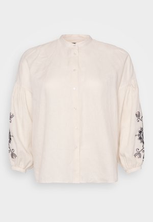 Camisa de lino marfil con cuello mandarín, largas mangas abullonadas con bordado floral negro en los puños y cierre frontal de botones.