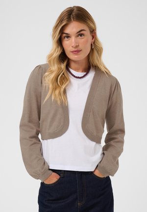 Jeune femme aux cheveux blonds ondulés portant un cardigan court beige sur une chemise blanche et un jean foncé, debout les mains dans les poches.