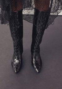 Stivali neri con tacco alto, con lacci a incrocio, punta appuntita, finitura lucida e lacci decorativi a croce che si estendono dalla caviglia a metà polpaccio.