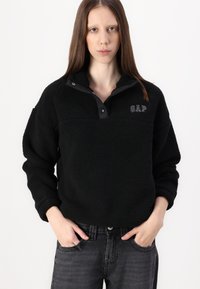 Zwarte fleece pullover met een korte kraag, halve knoopsluiting en geborduurd "GAP"-logo. Draagt het met donkergrijze jeans. Zachte textuur, relaxte pasvorm.
