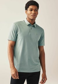 Camisa polo de color azul claro con cuello, mangas cortas, tapeta de tres botones y un pequeño logotipo bordado en el pecho.