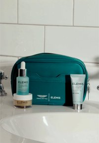 Trousse de maquillage teal avec fermeture éclair argentée ; contient trois produits de soins de la peau ELEMIS : sérum bleu avec compte-gouttes, baume nettoyant beige et crème blanche.