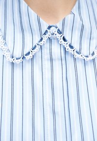 Γιακάς πουκάμισου με κάθετες ρίγες σε μπλε και λευκό, διακοσμημένος με λευκή βρόχο-κορδέλα κατά μήκος των άκρων, κουμπωμένος στο λαιμό.