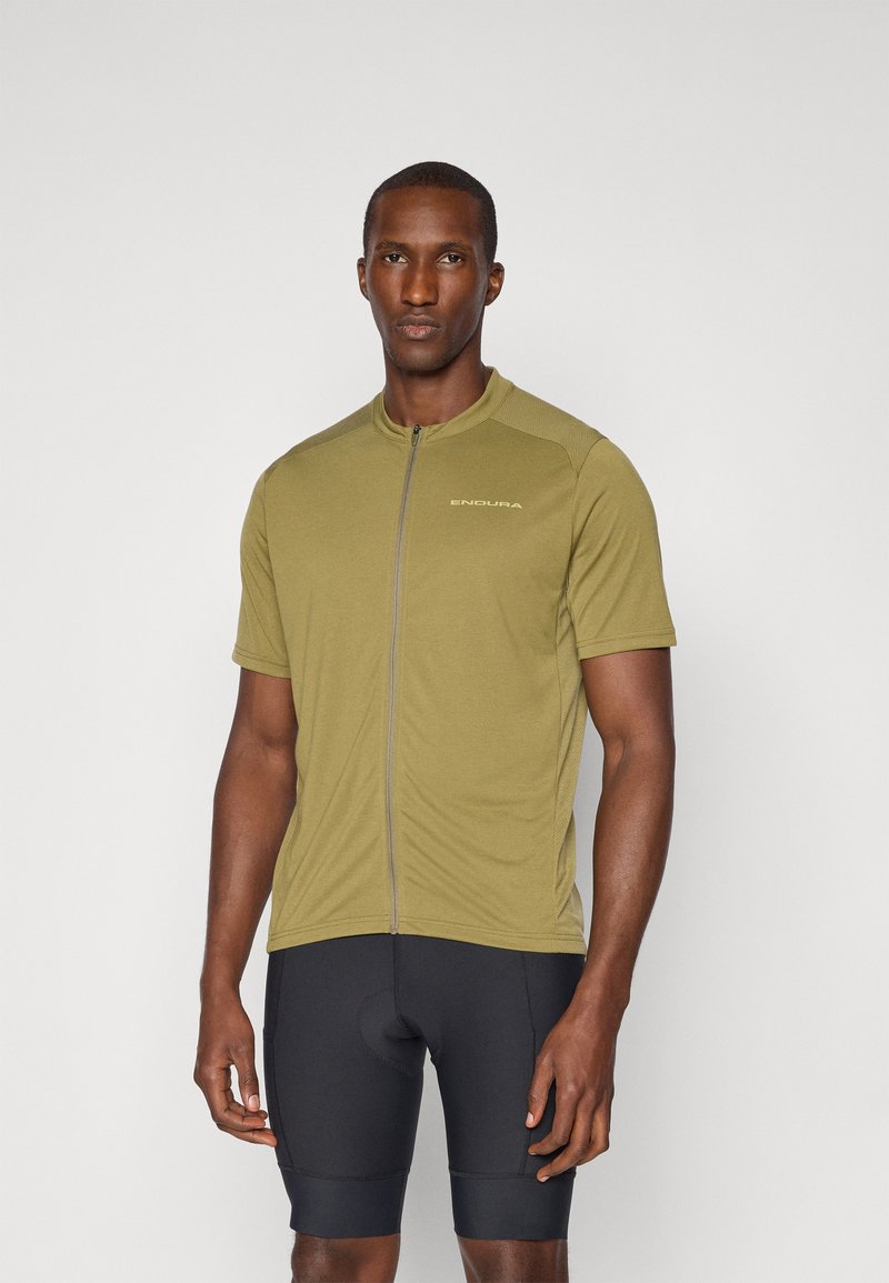 Olivgrünes Radtrikot mit Reißverschluss an der Vorderseite, kurzen Ärmeln und atmungsaktivem Stoff. Getragen mit schwarzen gepolsterten Radsportshorts.