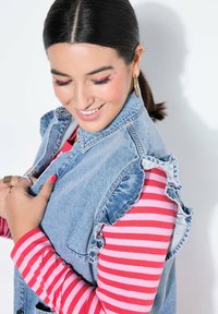 Spijkervest met ruches op de schouders, gedragen over een rood-roze gestreept shirt met lange mouwen. Met een zilveren ring aan de hand, roze nagellak en grote oorbellen.