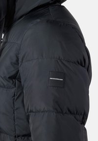 Schwarze Pufferjacke mit matter Oberfläche, ge quilteter Textur und einem quadratischen Logo-Patch mit der Aufschrift "SWEDEMOUNT" am linken Ärmel.