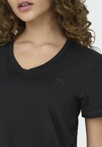 Zwarte V-hals T-shirt van een gladde stof, met een klein bordeauxrood rond logo op de linkerschouder. Korte mouwen, geen patronen.