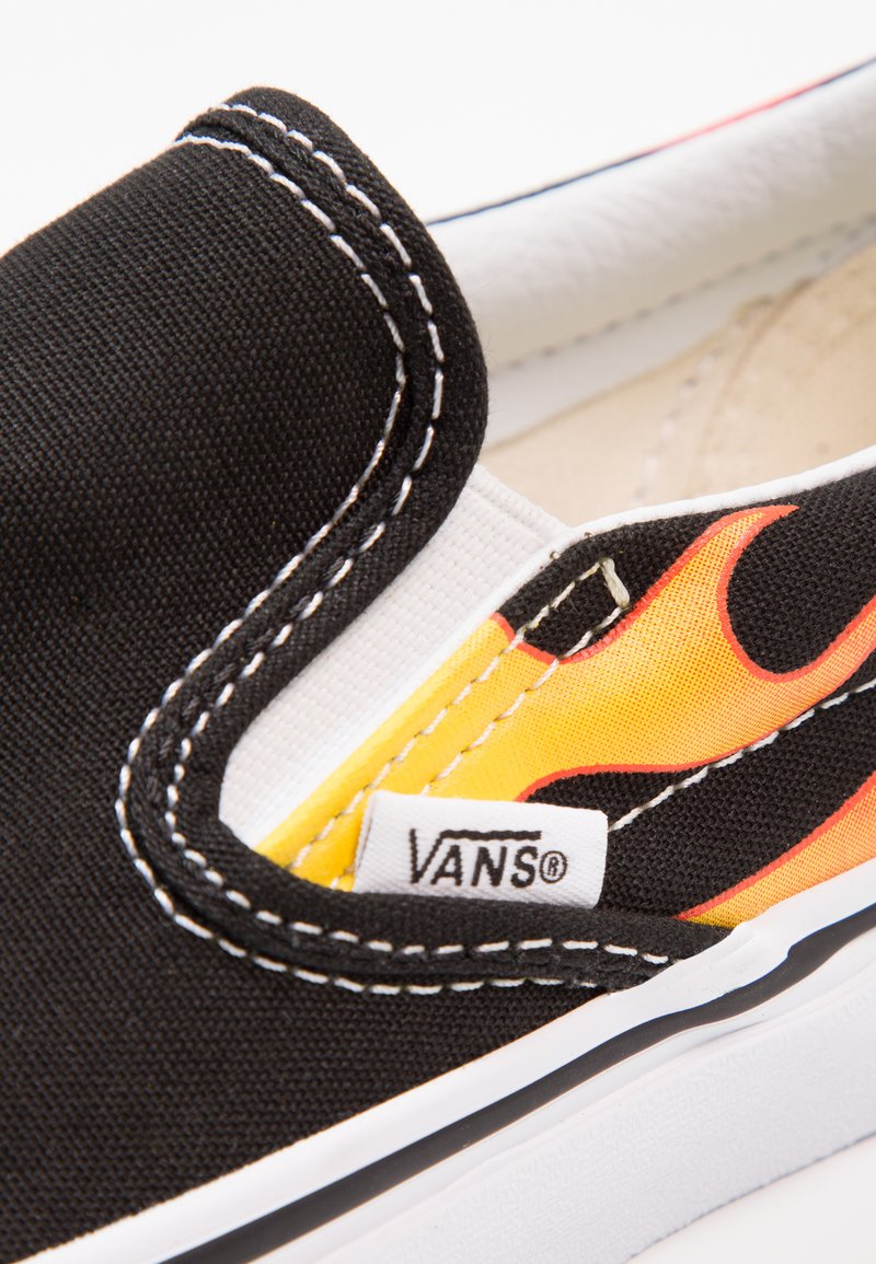 Zapato slip-on de lienzo negro con costuras blancas, diseño de llamas en amarillo y rojo, y una etiqueta de Vans en el lateral.